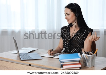 Woman in headset taking notes while passing online test via laptop at table indoors