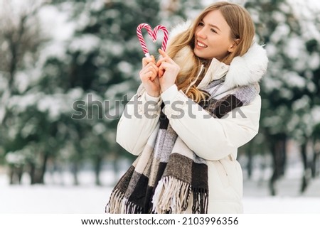 woman with heart shaped lollipop, happy woman in winter clothes, having fun with candy in her hands outdoors on a snowy day outdoors. Blonde girl with heart shaped lollipop, Valentine's Day concept