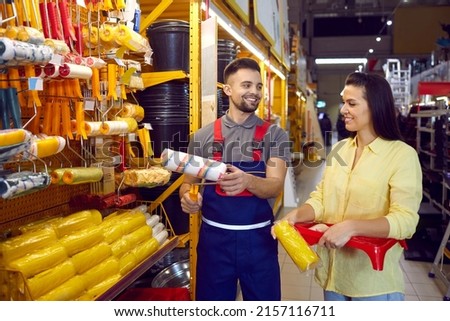 Woman with help of male salesman chooses supplies to repair house in building materials store. Friendly consultant in overalls helps female buyer to choose roller for painting walls. Repair concept.
