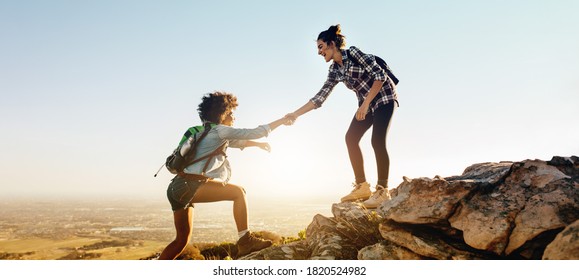 Woman Helping Her Friend To Climb The Cliff And Reach The Top Of Mountain. Friends Helping Each Other During Hiking A Mountain.