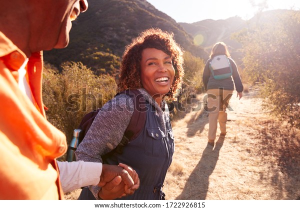 Woman Helping Man On Trail As Group\
Of Senior Friends Go Hiking In Countryside\
Together