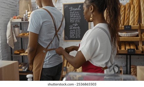 Woman helping man tie apron together in a cozy bakery room with shelves of bread and other baked goods in the background - Powered by Shutterstock