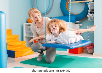 Woman helping a smiling boy to exercise on a therapy swing