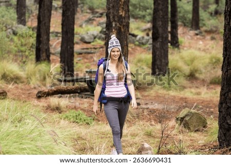 Woman in her 20s walking through pine forest clearing carrying purple backpack and wearing knit hat. Adventure, exploration, nature, outdoors, trekking, serenity, wilderness