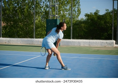 Woman in her 40s playing tennis on an outdoor court. She is in a ready position, preparing to serve the ball. The background features a fence and lush green trees, emphasizing a sunny, vibrant day. - Powered by Shutterstock