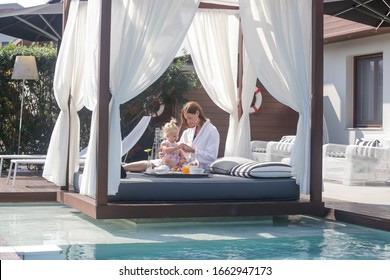Woman and her baby girl are enjoying breakfast in outdoor daybed with side curtains next to the swimming pool, the perfect beginning of summer day