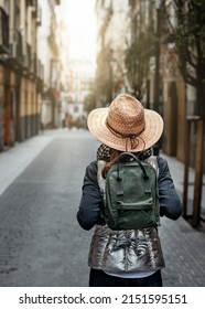 A Woman With Her Back Turned, Wearing A Hat And Backpack, Walking Down A Street In An Old Neighborhood In Madrid. City Tourism Concept.