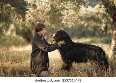 A woman and her Bernese Mountain Dog stand together in a sunlit meadow, surrounded by tall grass and trees. - Powered by Shutterstock