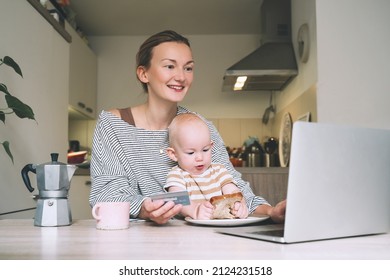 Woman With Her Child Buying Products Online Holding Credit Card In Hand In Kitchen At Home. Busy Mother With Laptop Paying For Shopping Online. Young Millennial Mom With Her Baby On Maternity Leave.