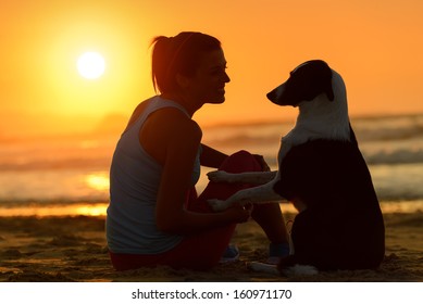 Woman With Her Cute Dog In The Beach On Golden Sunset Background. Girl Enjoying Her Pet Friendship And Affection Towards Beautiful Sun And Sea.