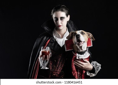Woman and her dog in similar vampire costumes for halloween. Young female with glass of red drink and her pet puppy dressed up in same Dracula costume