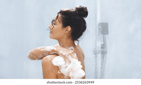 A woman with her hair in a bun enjoys a refreshing shower, using a lathered loofah to gently cleanse her skin. Her relaxed expression and the calming atmosphere make this image ideal for promoting - Powered by Shutterstock