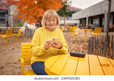 Woman in her late sixties sits in the street cafe in autumn, surrounded by fall colors. She skillfully uses her smartphone. Adaptability in the digital era, a tech-savvy senior and modern technology - Powered by Shutterstock