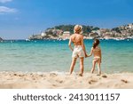 Woman and her little daughter in swimsuit standing together on the beach, watching at the Port de Soller bay in Mallorca. Holidays with children concept.