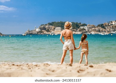 Woman and her little daughter in swimsuit standing together on the beach, watching at the Port de Soller bay in Mallorca. Holidays with children concept.