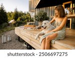 A woman and her loyal dog enjoy a serene evening on a wooden terrace, surrounded by nature. The soft light of the setting sun enhances the peaceful and joyful moment shared between them