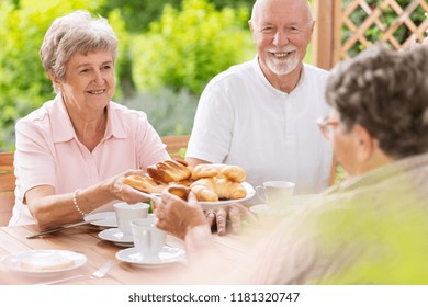 A Woman In Her Old Age Handing Pastry To Her Colleague Friend During An Afternoon Snack Time On A Patio Of A Happy Nursing Home.