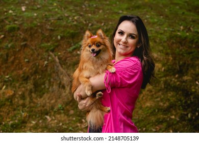 Woman With Her Pet Dog On Her Lap. The Brazilian Wearing A Pink Blouse And The Dog With A Bow On Her Head.