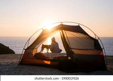 Woman And Her Pet Dog Sit At Beach Next To Tent, Solitude Camping In Nature. Sunset Shot Of Young Woman Have Self Exploration Time And Connection To Nature. Tranquility And Mindfulness