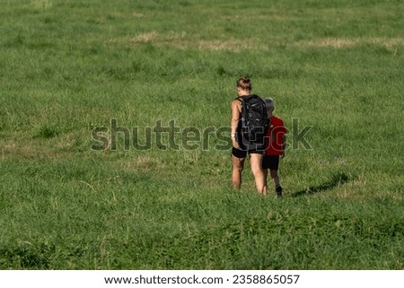 A woman and her son are exploring a green field on a summer day. They wear backpacks and walk among the tall grass. The sun illuminates them brightly. Concept of natural beauty and family adventures