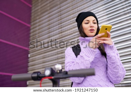 Woman in her twenties with electric scooter recording voice note with a smartphone outdoors. Lifestyle concept.
