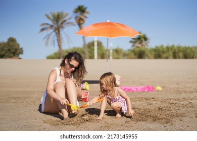 A Woman And Her Young Daughter Playing With The Sand To Bury Their Feet On The Beach