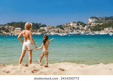 A woman and her young daughter in swimsuits standing on the beach, observing Port de Soller bay in Mallorca, enjoying a sunny day together - Powered by Shutterstock