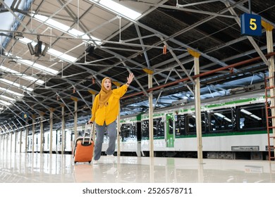 Woman in Hijab Waving at Train Station Platform. Smiling woman in a yellow hijab waves while running at a busy train station platform with her luggage. She appears excited and ready for travelling. - Powered by Shutterstock