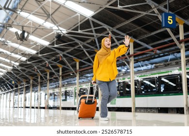 Woman in Hijab Waving at Train Station Platform. Smiling woman in a yellow hijab waves while running at a busy train station platform with her luggage. She appears excited and ready for travelling. - Powered by Shutterstock