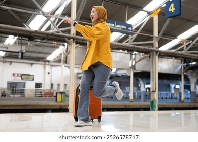 Woman in Hijab Waving at Train Station Platform. Smiling woman in a yellow hijab waves while running at a busy train station platform with her luggage. She appears excited and ready for travelling. - Powered by Shutterstock