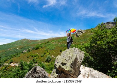 A Woman Hiker With A Backpack, Perched On A Rock At The Top Of A Mountain, Arms Raised Holding A Rainbow Flag Fluttering In The Wind. With A Blue Sky And Mountainside In The Background.