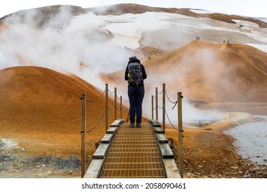 Woman hiker with a backpack walking over a small wood bridge at Hveradali geothermal area in Kerlingarfjoll mountain. Snowfields and steaming fumaroles are visible in the background. Iceland Highlands