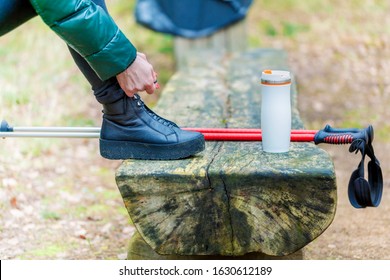 Woman hiker with trekking sticks near bench fixing her boot in forest
