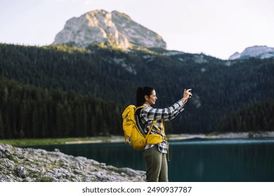 A woman hiker with a yellow backpack, smiling and taking a photo of the scenic mountain lake with her smartphone. - Powered by Shutterstock