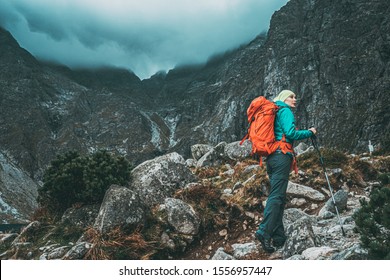Woman Hiking in High Mountains on Stormy Moody Weather. Female tourist with backpack on a trail with clouds and fog covered mountains background, spring, autumn or winter. Extreme Adventures.  