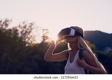 Woman Hiking At Sunrise Using VR Goggles To Explore The Metaverse. Blond Woman On Holiday Hiking In A Forest Using VR Goggles To Experience Augmented Virtual Reality