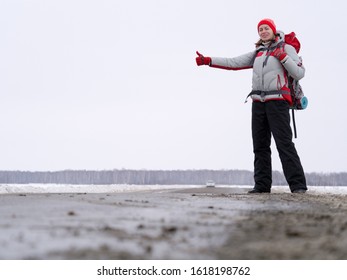 Woman Hitch Hiker Try To Catch A Car Showing Thumb Up On A Road To Share A Ride. Winter Time, Front View, Selected Focus. Big Backpack And Warm Sportawear. Adventure, Activity, Good Fortune, Freedom