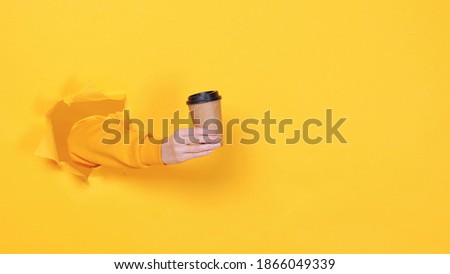 Woman hold in hand paper cup of coffee or tea isolated through torn yellow wall background studio. Copy space advertisement place for text image promotional content Advertising area workspace mock up