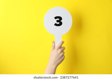 Woman holding auction paddle with number 3 on yellow background, closeup