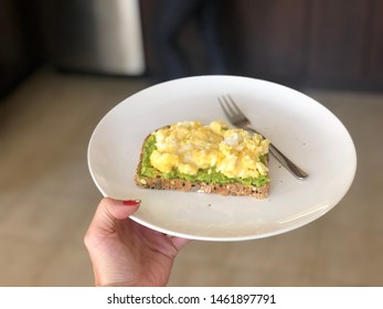 Woman Holding Avocado Toast With Scrambled Eggs And Crushed Black Pepper On White Plate Held In Hand Top View Healthy Breakfast In The Morning