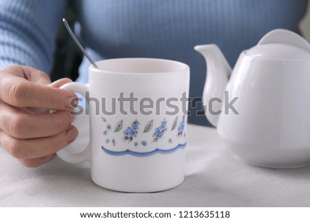 Woman holding beautiful mug with spoon and blurred white porcelain teapot on background