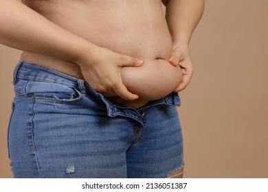 Woman holding big belly with hands showing fat in blue jeans on beige background. Sudden weight gain. Visceral fat. Body positive. Tight little clothes. Need for wardrobe change.