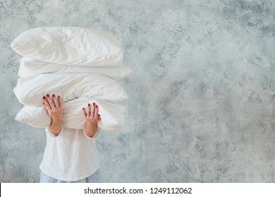 Woman holding big pile of white soft cozy pillows on gray background. bedding and sleeping concept