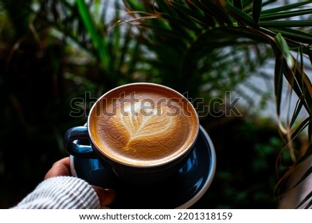 woman holding Black cup of cappuccino with latte art at outdoor cafe. tropical plant.