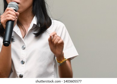 The Woman Is Holding The Black Microphone In Her Right Hand And Do The Public Speaking On The Stage With White Background, In Concept Of Presentation, Talk Show, Confidence.