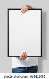 Woman Holding A Blank Poster With Black Frame Mockup Isolated On A Gray Background. 