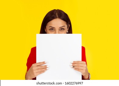 Woman holding a board paper hiding her smile behind it. Mixed race model isolated on yellow background with copy space. Horizontal image. Natural, no makeup.