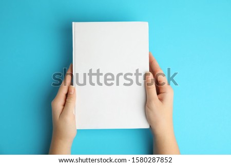 Woman holding book with blank cover on blue background, top view