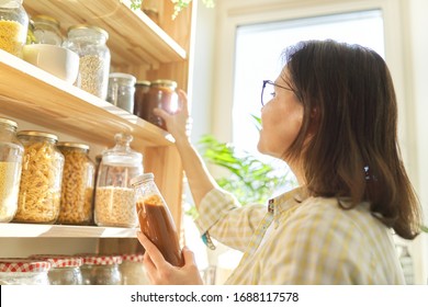 Woman holding bottle with ketchup, picking food from storage cabinet in kitchen, storage with wooden shelves