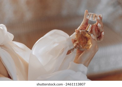 A woman is holding a bottle of perfume. The bottle is made of glass and has a gold cap. The woman is wearing a white blouse and is holding the bottle in her hand - Powered by Shutterstock